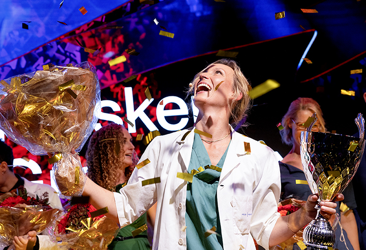 En person i hvit frakk og grønne klær holder en stor blomsterbukett og et trofé. Rundt personen faller det konfetti, og andre personer med blomsterbuketter er delvis synlige i bakgrunnen.