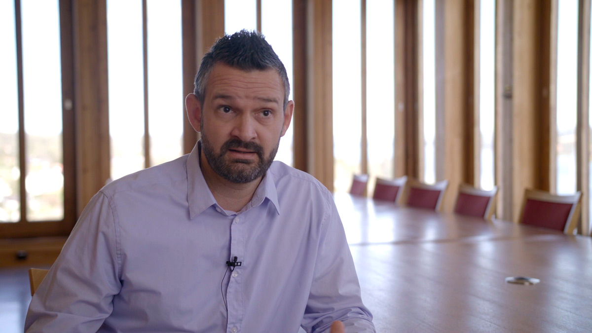 Mann med mørkt hår, kort skjegg  og blå skjorte sitter i et tomt møterom og snakker mot kamera.